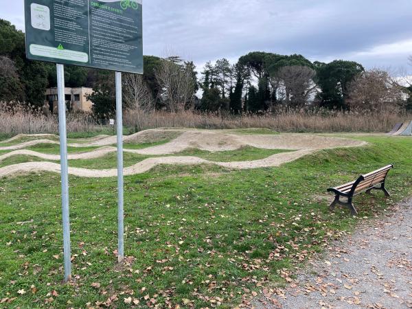 Bike Pump Track dans une zone herbeuse. A gauche, un panneau de signalisation. A droite, un banc.